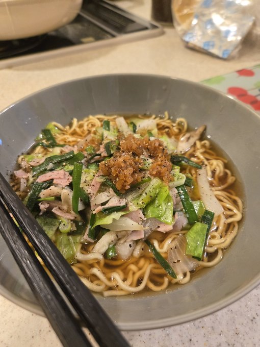 The image features a bowl of noodles with various toppings, including meat, vegetables, and cheese. The bowl has a gray color and is placed on top of a table or countertop. There are chopsticks resting next to the bowl, suggesting that someone was eating from it earlier.
