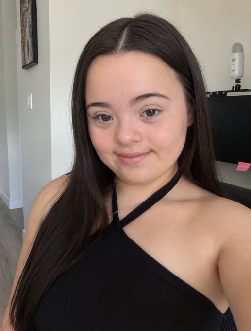 A young woman with long brown hair smiles slightly while wearing a black halter top, with what appears to be a Blue Yeti microphone visible in the background. The image relates to the post's humor about appearing cheerful on the outside while struggling with Bitcoin's volatile price drops—a relatable "fake it till you make it" crypto investor expression.