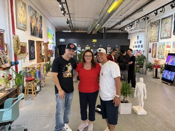 Three people pose together inside an art gallery filled with paintings, sculptures, and decorative pieces, with the woman in red standing between two men in casual attire.