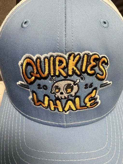 A close-up of a light blue baseball cap featuring a colorful embroidered patch that reads 'QUIRKIES WHALE 2026' with a cartoon skull design.