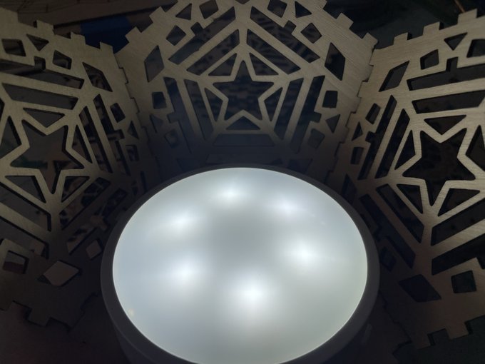 An overhead view of an illuminated round LED light fixture surrounded by laser-cut wooden panels featuring intricate geometric and star-shaped cutout designs.