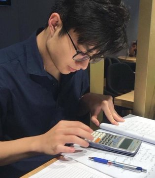 A young man with glasses using a scientific calculator while writing in a notebook at a desk.