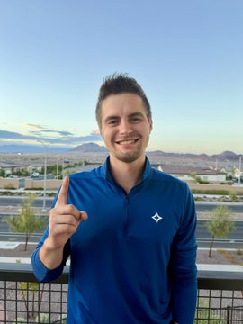 A man in a blue pullover (featuring the CleanSpark diamond logo on the chest) smiles at the camera while pointing upward, standing on a balcony with mountains visible in the background — celebrating his one-year anniversary at CleanSpark.