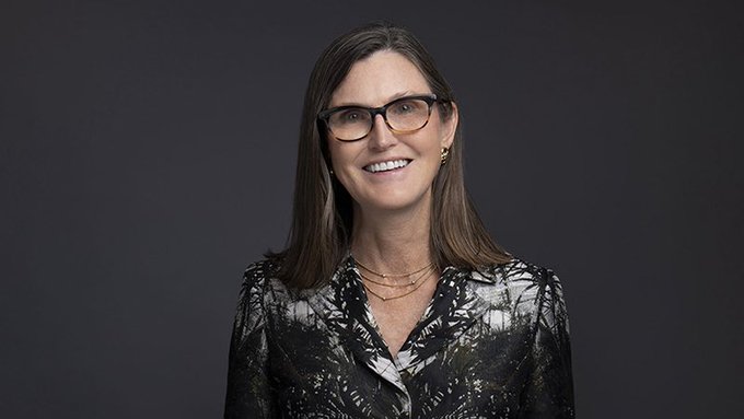 A headshot of a smiling woman with glasses and shoulder-length brown hair wearing a patterned blouse against a dark background.