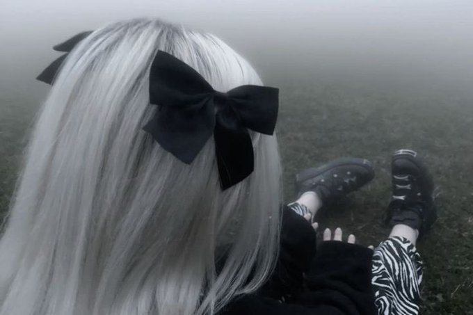 A person with long silver hair adorned with a large black bow, seen from behind sitting in a foggy field, wearing zebra-print pants and black boots.