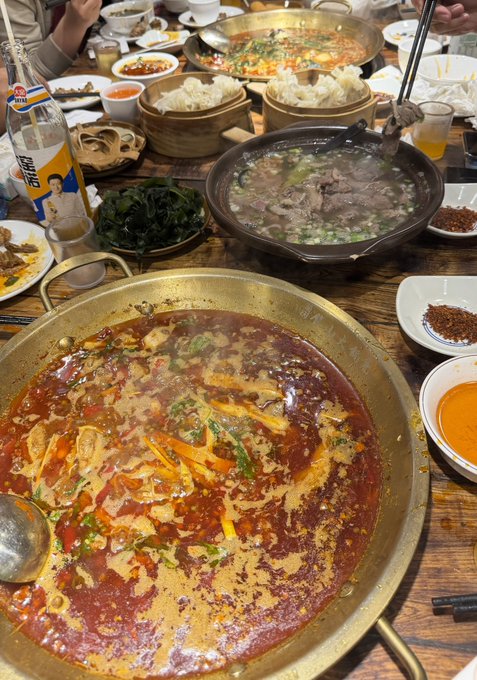 A table filled with Chinese hot pot dishes, including a large pan of spicy red broth, a bowl of beef soup, bamboo steamers with buns, and a bottle of Snow (雪花) beer.