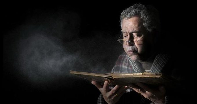 A thoughtful older man with glasses intently reads a worn, ancient book in dramatic low light, dust particles floating through a beam of light above the pages—evoking the forgotten, dusty relics of memory referenced in the post about "1 billion."