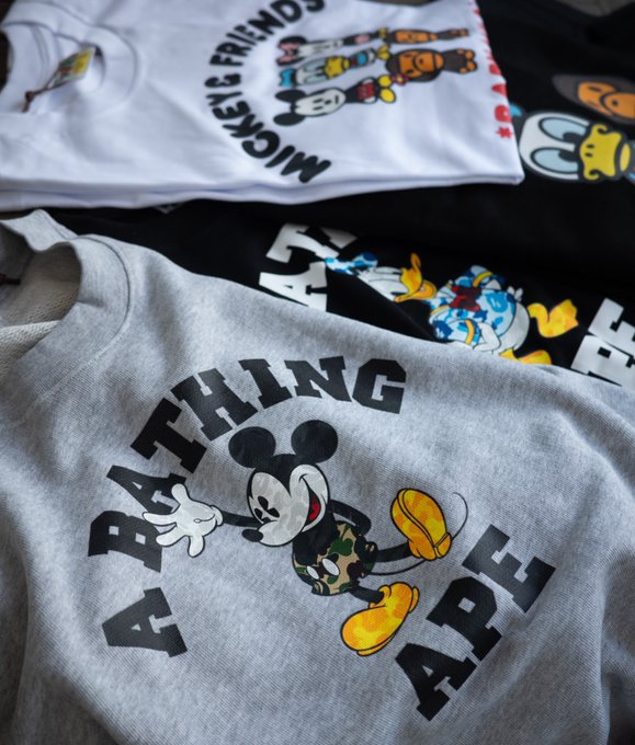 A photograph of stacked t-shirts featuring Disney characters, with a gray shirt in the foreground reading 'A BATHING APE' around an image of Mickey Mouse wearing camouflage pants.