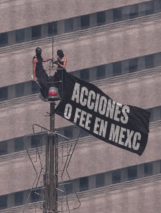 Two masked protesters sit atop a pole near a building, unfurling a large black banner reading 'ACCIONES O FEE EN MEXC' as part of an activist demonstration.