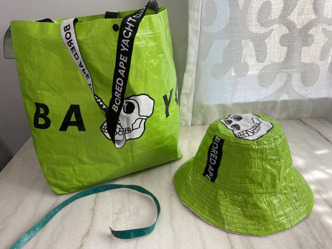 A bright green Bored Ape Yacht Club (BAY) branded tote bag and matching bucket hat with skull logo, displayed alongside a measuring tape on a marble surface.