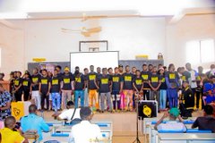 A group photo of people, possibly students or participants, posing together in front of a sign while holding yellow papers, with audience members seated in the foreground.