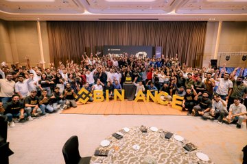 A large group of attendees gathers at a Binance event, holding yellow letters spelling "BINANCE," reflecting the post's theme of one centralized exchange fostering numerous decentralized crypto communities.