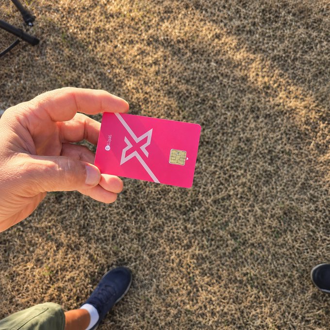 A hand holds a bright pink Crypto.com card featuring a chip and a stylized "X" logo, displayed over dry grass with feet in black sneakers visible below. The scene captures the summer-ready, on-the-go vibe of the post, showing the card as a portable cold wallet suited for outdoor, travel-friendly use.