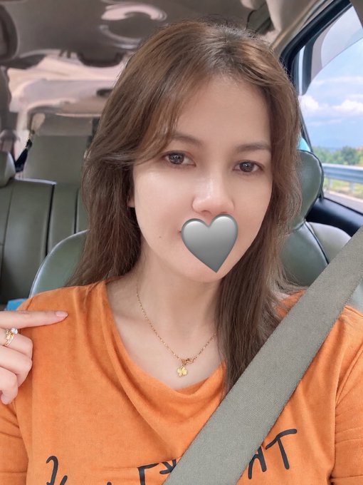 A woman takes a selfie in a car wearing an orange t-shirt and gold necklace, with a heart emoji sticker covering her mouth.