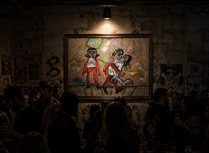 A dimly lit bar or venue where a crowd of people is gathered beneath a framed artwork depicting cartoon monkeys in orange jumpsuits.