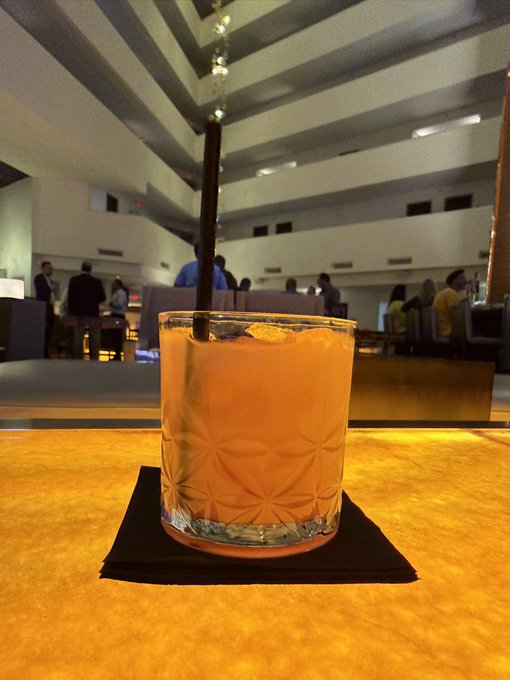 An orange-colored cocktail served in a rocks glass on a black napkin at a bar, with a spacious multi-level interior visible in the background.