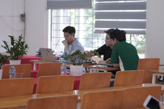 Three young men sitting at desks in a classroom or study area, with one working on a laptop and water bottles and small potted plants on the wooden desks.