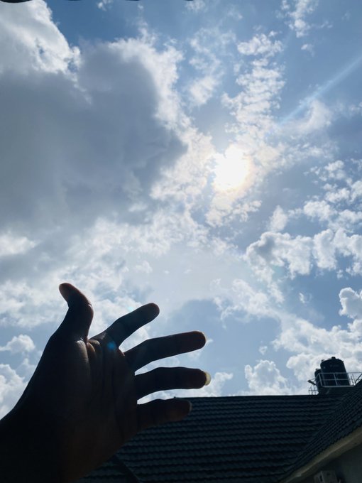A silhouetted hand reaching toward a bright sun in a partly cloudy blue sky, with a tiled rooftop and water tower visible in the lower right corner.