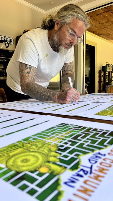 A man with long gray hair, beard, glasses, and heavily tattooed arms signing prints with a white marker at a table displaying colorful graphic artworks featuring green geometric patterns and a rabbit face.