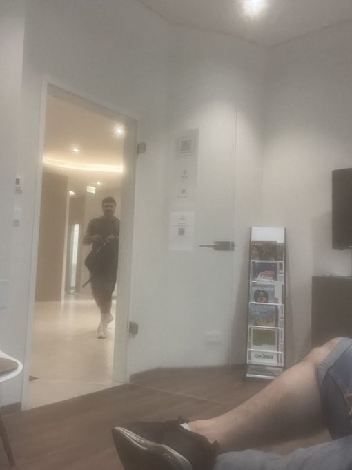 A casual interior shot showing a person's legs in the foreground while another person carrying a bag stands in the doorway of a modern, minimalist apartment.