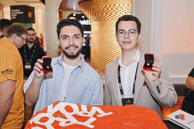 The image shows a group of people standing around, with two men holding up their engagement rings for the camera. The man on the left is wearing an orange shirt while the other man has his ring displayed on his finger. They are surrounded by several other people in various positions within the scene. In addition to the main subjects, there is also a book visible in the image, which could be related to their engagement or simply part of the background setting.