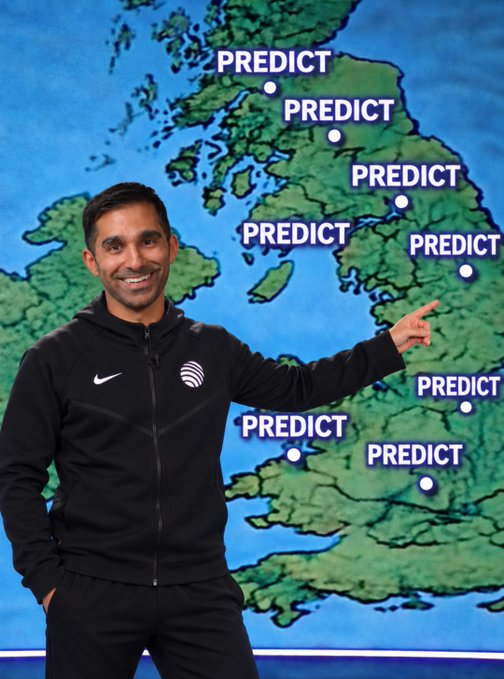 A man in a black tracksuit smiling and pointing at a weather map of the UK and Ireland, which is covered with multiple 'PREDICT' labels over various locations.