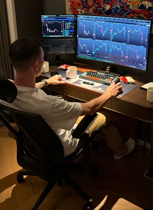 A person sitting at a desk trading setup with multiple monitors displaying candlestick charts, wearing headphones around their neck while operating a keyboard and mouse.