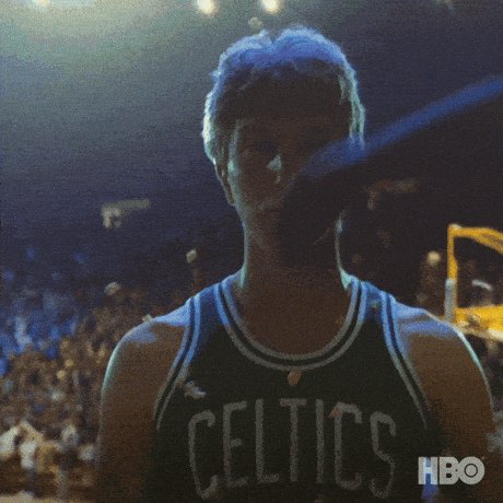 The image features a man wearing a green and white jersey with the word "Celtics" written across his chest, standing in front of an audience at a basketball game. The crowd behind him appears to be engaged in watching the game, as indicated by their attentive expressions. A baseball bat is visible near the top right corner of the image, suggesting that this might be a sports event or a game with multiple sports elements combined into one.