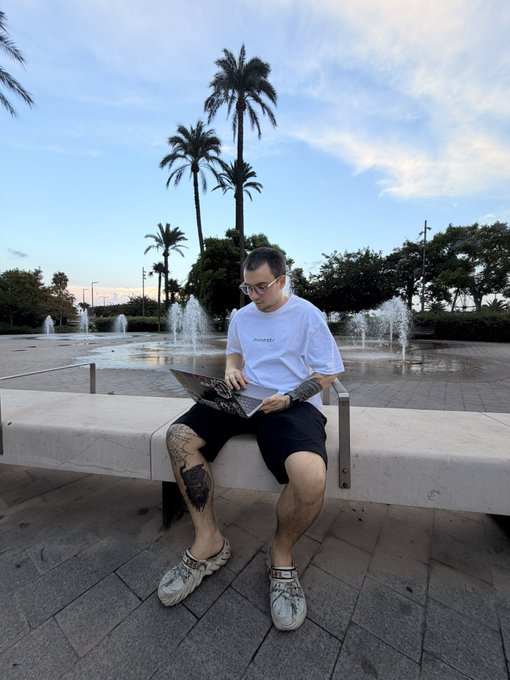 A man wearing glasses, a white t-shirt, and black shorts sitting on a stone bench working on a laptop in a plaza with fountains and palm trees.