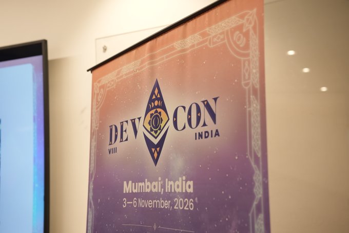 A purple and orange banner for 'Devcon VIII India' in Mumbai, dated 3-6 November, 2026, hangs indoors near a screen.