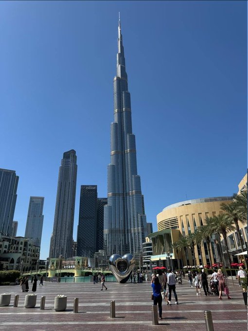 A daytime photograph of the Burj Khalifa in Dubai, the world's tallest building, towering over a plaza with pedestrians and surrounding skyscrapers.