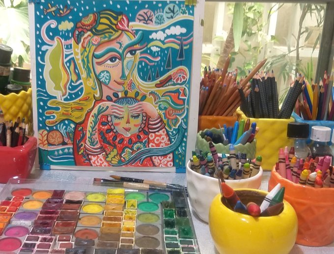 An artist's creative workspace featuring a vibrant, colorful painting of two stylized figures, surrounded by containers full of colored pencils, crayons, and an open watercolor palette.