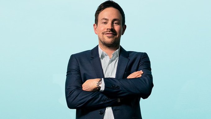 The image features a man dressed in a suit with his arms crossed, looking directly at the camera. The background of the photo has a light blue color that contrasts nicely with the man's dark-colored suit. The man appears confident and composed, and there is also some text within the image that seems to be related to investing or financial news, but it does not provide much additional information about him beyond his attire.