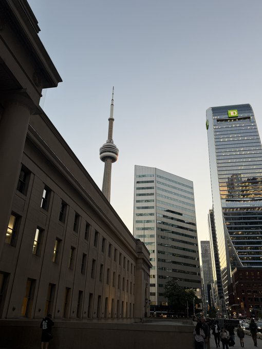 An urban street scene at dusk in Toronto, featuring the historic stone facade of a building in the foreground, the CN Tower rising in the distance, and the glass TD Canada Trust Tower alongside other modern skyscrapers.