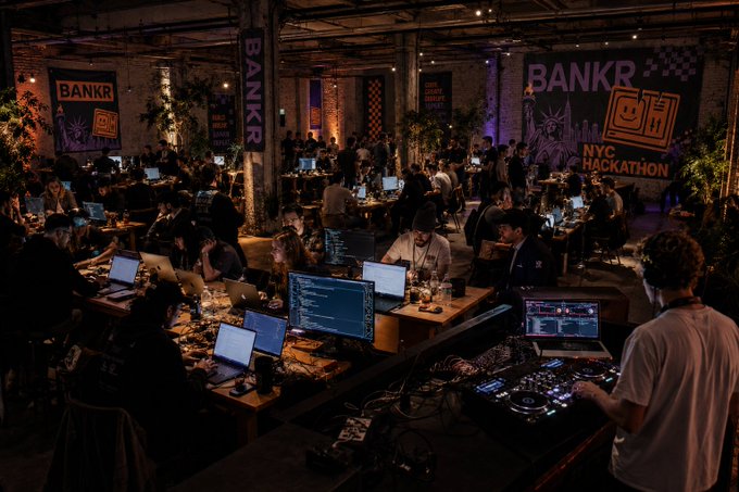 The image shows a large room filled with people working on their laptops, likely participating in an event or competition related to technology or programming. The room has multiple tables covered with laptops and computer equipment, indicating that this gathering might be focused on collaborative work or problem-solving activities. A DJ is also present in the scene, suggesting that there could be a music-related aspect to the event as well. The people are sitting at various tables, some of them working closely together while others have their laptops open and facing away from each other. Overall, it appears to be an environment where individuals can come together to share ideas or work on projects in a collaborative setting.
