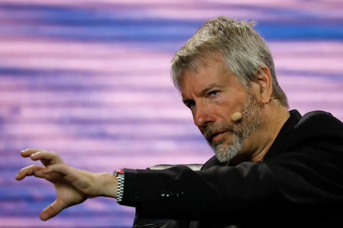 A man with gray hair and a beard, wearing a dark suit and a headset microphone, gestures with his hand extended while speaking on stage.
