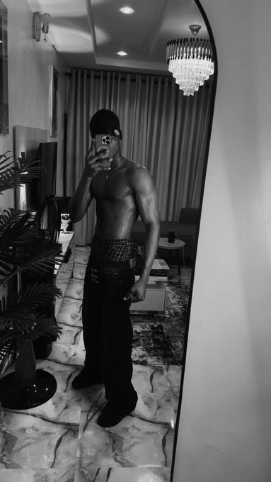 A black-and-white mirror selfie of a shirtless man in a beanie holding a smoking object, standing in a room with a chandelier and marble flooring.