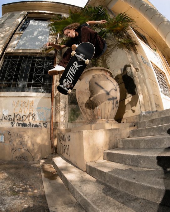 A young male skateboarder performs a trick, jumping over a stone railing next to a building with graffiti and a large decorative urn.