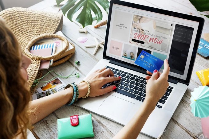 A woman shopping online using a laptop and credit card, with the screen displaying an e-commerce website featuring 'SHOP NOW' promotional banners.