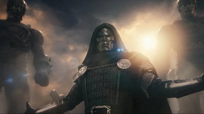 A dark, dramatic scene of a hooded masked figure in armor with arms outstretched, flanked by two shadowy robotic figures with glowing green eyes against a hazy golden background.