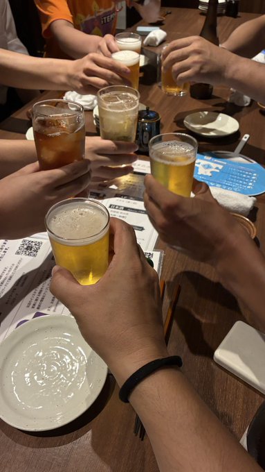 A group of people clinks glasses filled with beer at a restaurant table, with one visible coaster featuring a QR code, capturing a toast moment that reflects the post's description of a STEPN Aji Fry Club gathering in Kayabacho.