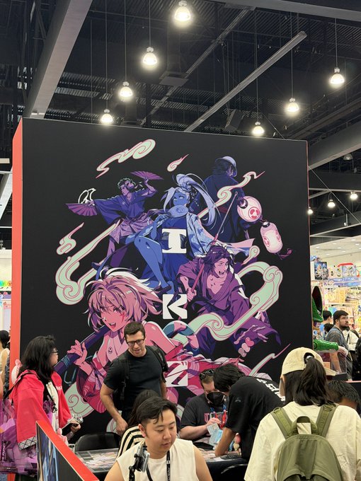 An anime convention booth featuring a large vertical banner with multiple illustrated characters, surrounded by attendees and vendors.