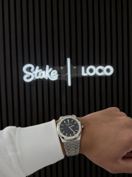 A close-up of a wrist wearing an Audemars Piguet Royal Oak watch, posed in front of a black wall with illuminated 'Stake | LOCO' signage.