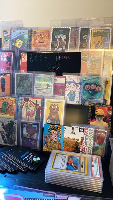 A display of trading cards features a chalkboard sign reading 'BEST PHCDS ENJOY', showcasing various illustrated cards and a stack of cards with a 'TWWING' character.