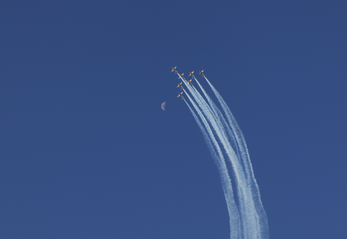 Five jets in formation flying upward against a deep blue sky, leaving long white vapor trails, with a faint crescent moon visible in the background.