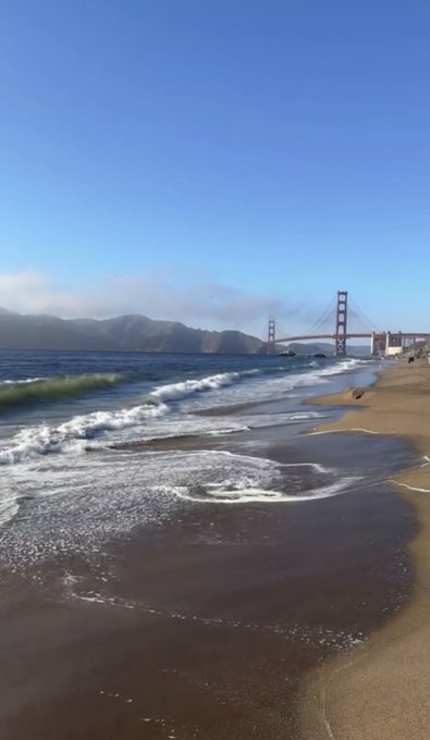 A scenic view of the Golden Gate Bridge seen from a sandy beach with ocean waves rolling in under a clear blue sky.