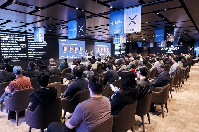A large conference hall with a seated audience facing a stage, with blue banners reading 'ripple' and 'XRP TOKYO' hanging from the ceiling and screens displaying 'XRP TOKYO' and 'Ripple' branding.