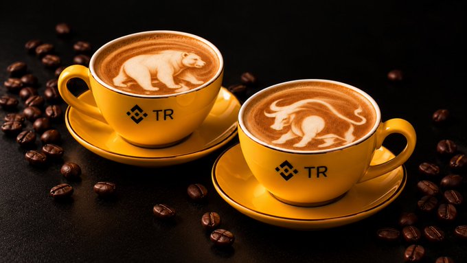 Two yellow espresso cups branded with "TR" feature intricate latte art of a bear and a bull, surrounded by scattered coffee beans, symbolizing the bear and bull market sentiments on World Coffee Day.