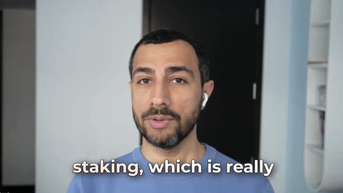 The image features a man wearing a blue shirt with white text that reads "staking, which is really". The man appears to be looking directly at the camera while speaking into an unseen microphone. The background of the image shows a room with a door visible in the scene.