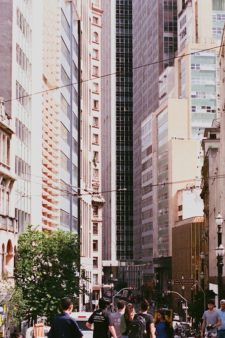 A bustling city street scene features towering skyscrapers and pedestrians walking along the sidewalk under overhead cables.